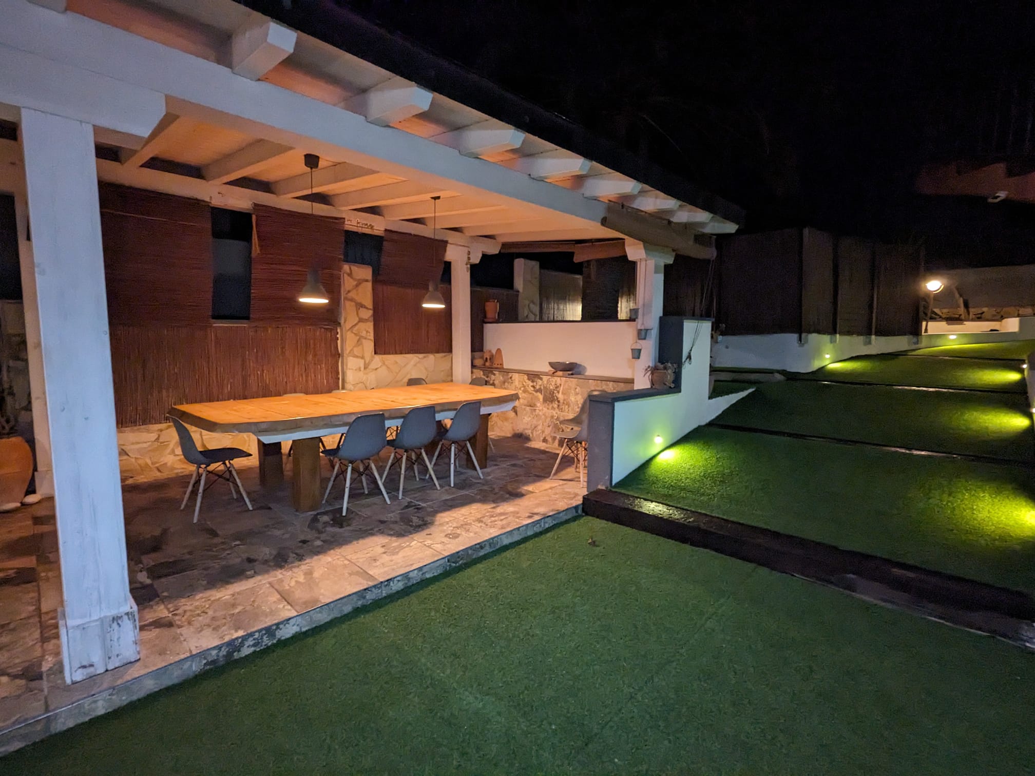 Comedor exterior bajo pérgola de madera blanca con mesa de madera maciza, sillas modernas y suelo de piedra, junto a un jardín iluminado con césped artificial.