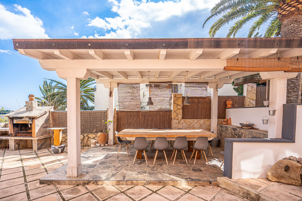 Comedor exterior bajo una pérgola de madera blanca con mesa robusta, sillas de diseño y zona de barbacoa de piedra integrada en el patio pavimentado.