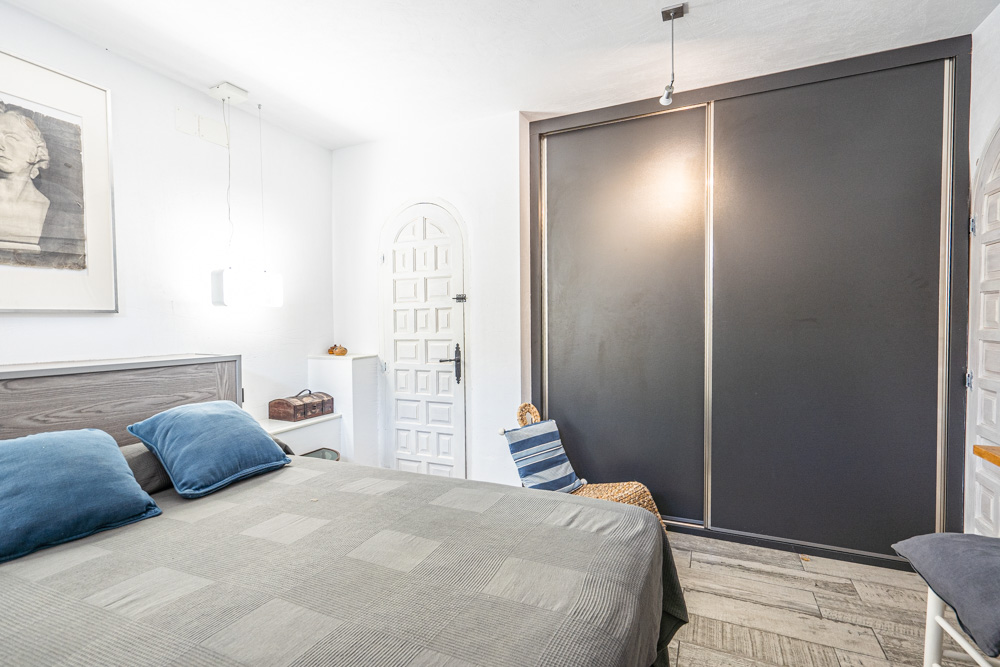 Dormitorio con armario empotrado de puertas correderas oscuras, suelos de gres efecto madera y una puerta de paso blanca con diseño de arco tradicional.