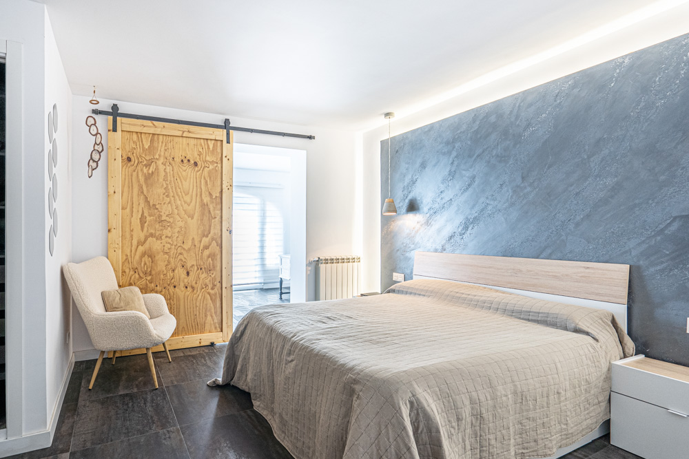 Dormitorio con pared de acento gris texturizada, iluminación LED indirecta, suelos de gres oscuro y una puerta corredera de madera natural sobre riel negro.