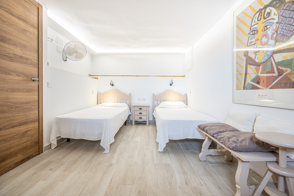 Dormitorio luminoso con dos camas individuales, suelos cerámicos de efecto madera, iluminación LED indirecta en el techo y ventilador de pared.