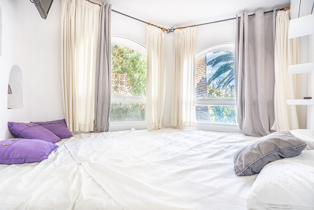 Dormitorio luminoso con ventanas de arco que ofrecen vistas al jardín mediterráneo. Incluye textiles en tonos blancos y violetas, y aire acondicionado.