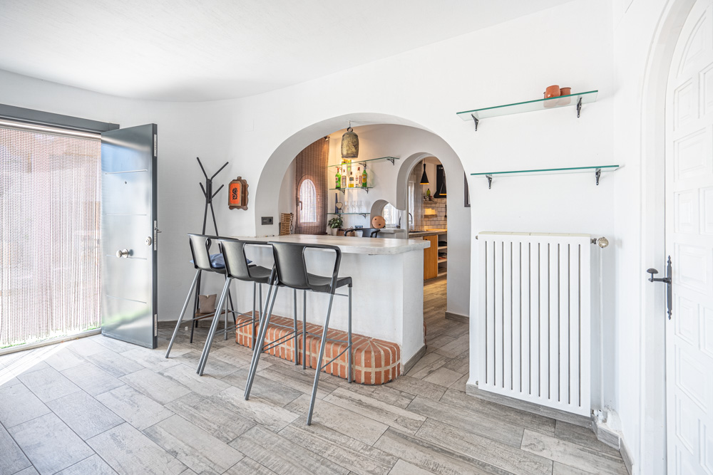 Entrada con barra de desayuno integrada, suelos de gres efecto madera y un arco decorativo que conecta con la cocina. Incluye radiador y acceso directo al exterior.