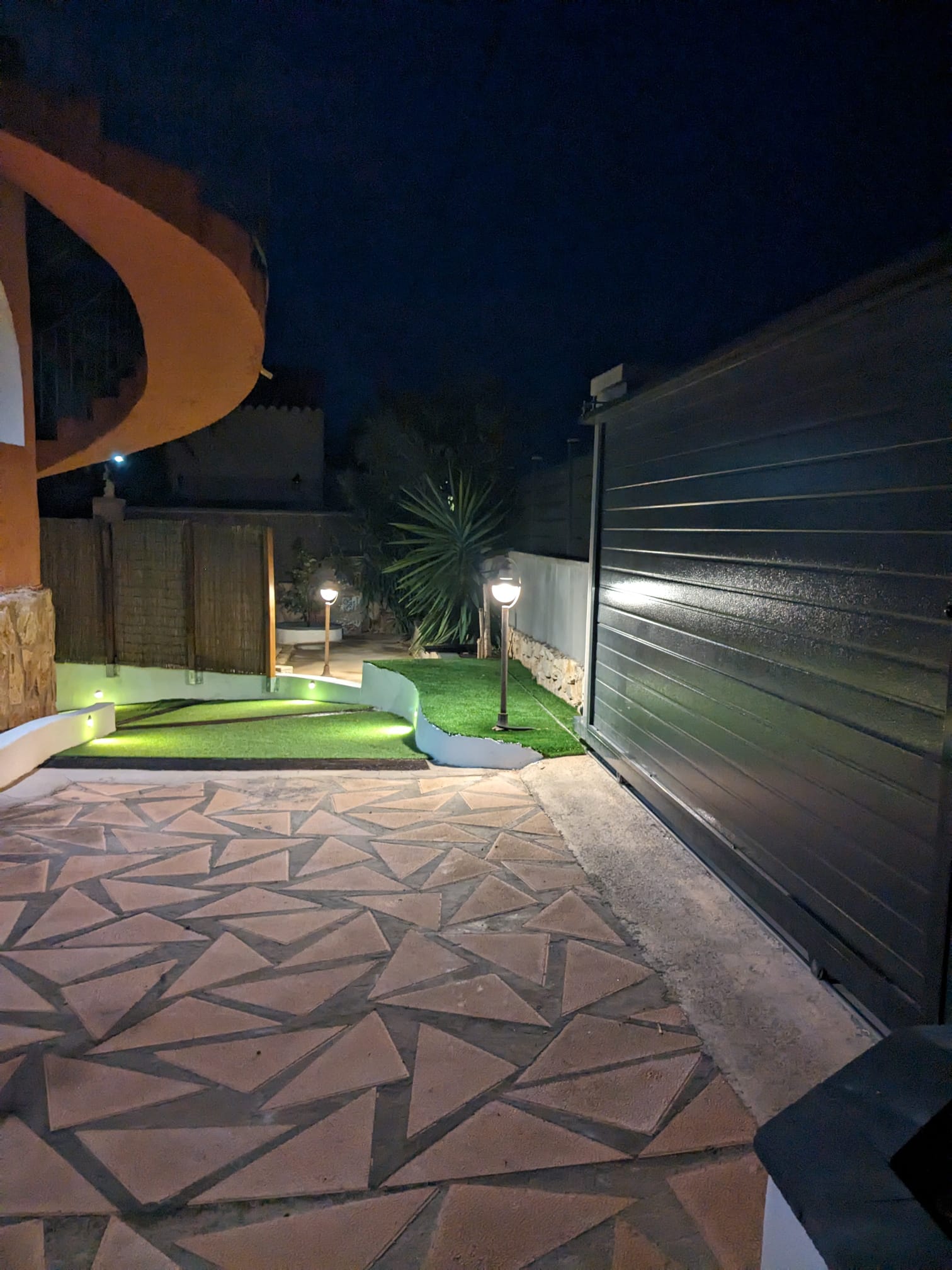 Entrada exterior nocturna con portón corredero gris, pavimento de piedra geométrica e iluminación integrada en muros y zonas de césped artificial.