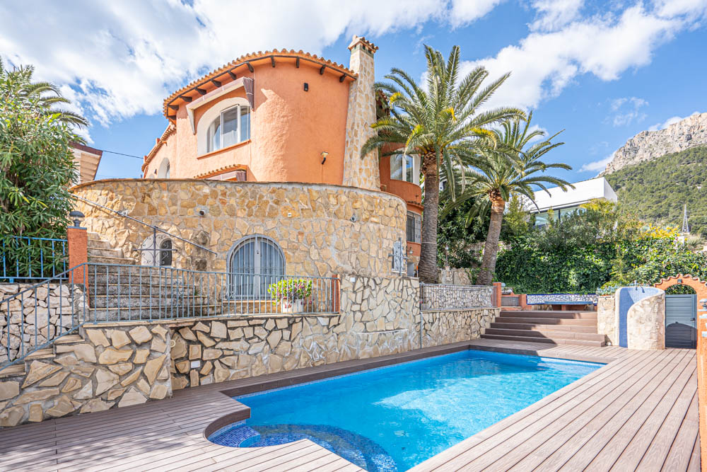 Exterior de la villa con piscina privada, rodeada de una terraza de tarima y muros de piedra natural. Se aprecia la fachada en tono terracota junto a palmeras y vistas a la montaña.
