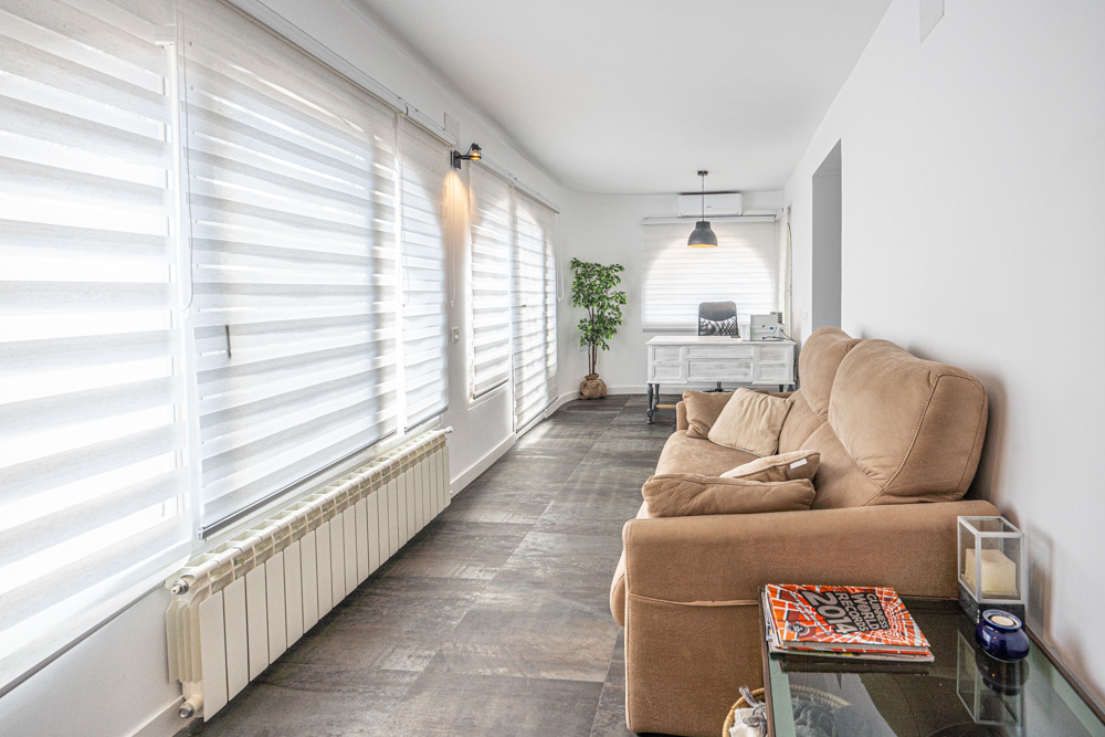 Naya luminosa con suelo de gres oscuro, zona de estar con sofá beige y rincón de oficina. Incluye radiador, aire acondicionado y amplios ventanales con persianas de lamas blancas.