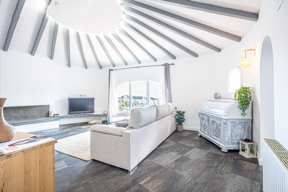 Salón circular con vigas de madera radiales en gris, suelos de gres oscuro y ventanal con vistas al entorno urbano de Calpe.