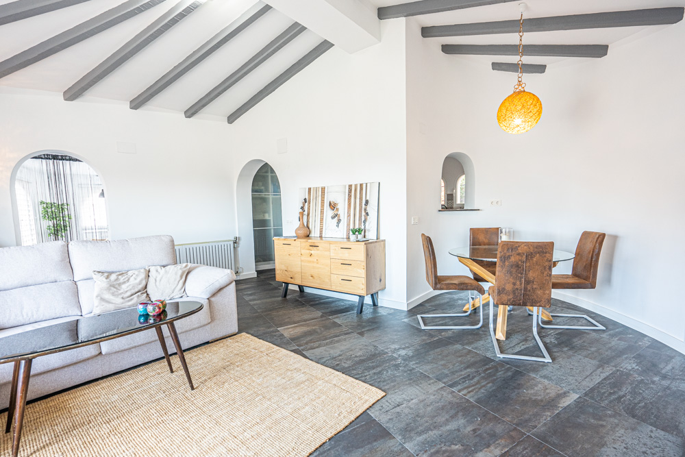Salón comedor con techos de vigas de madera gris, suelos de gres oscuro y arcos arquitectónicos que conectan con la cocina y la naya exterior.