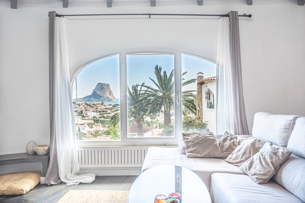 Salón luminoso con un gran ventanal de arco que enmarca vistas directas al Peñón de Ifach y al mar Mediterráneo, complementado con techos de vigas de madera.