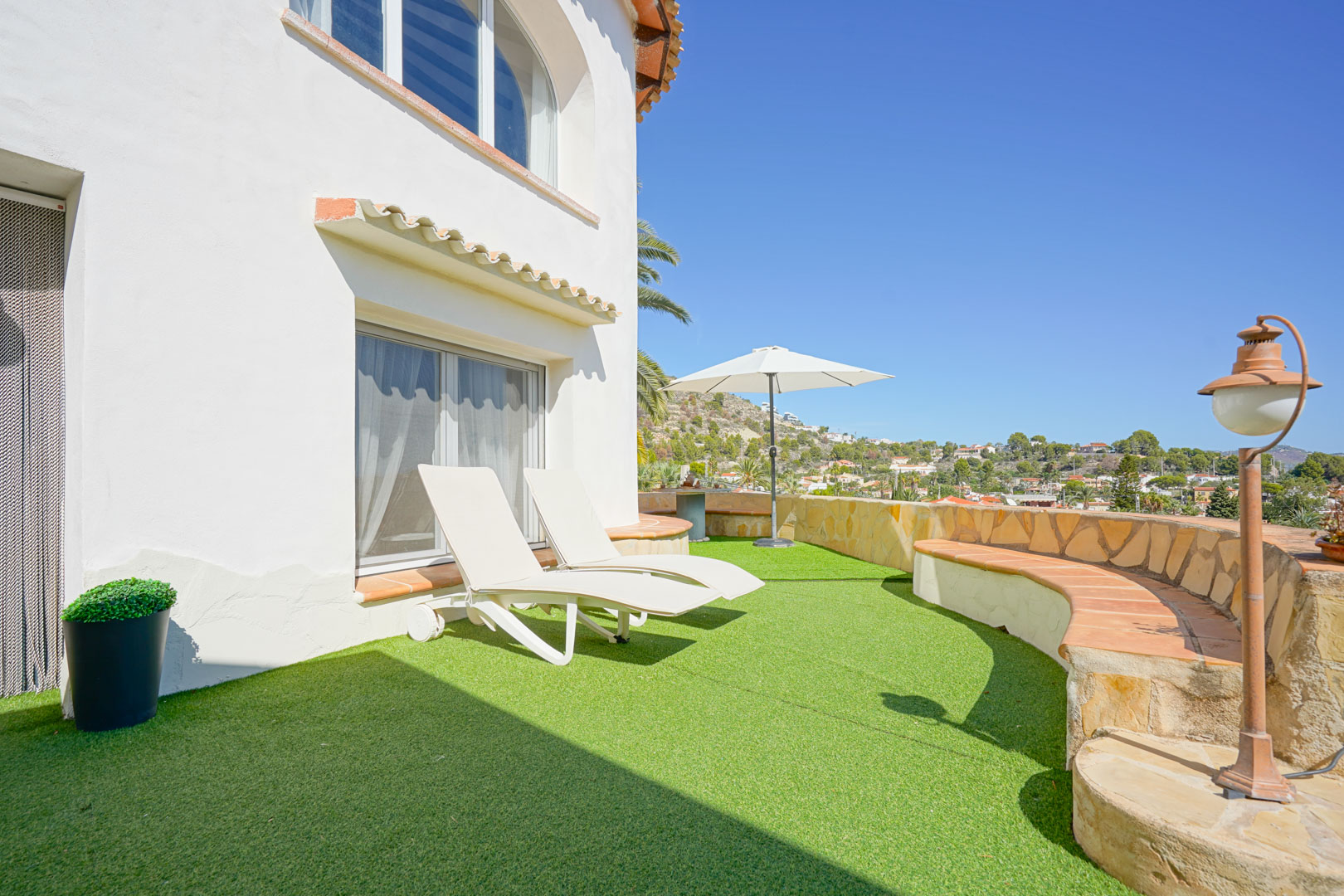Terraza con césped artificial, tumbonas blancas y un banco curvo de piedra con vistas despejadas a los alrededores de Calpe bajo un cielo despejado.