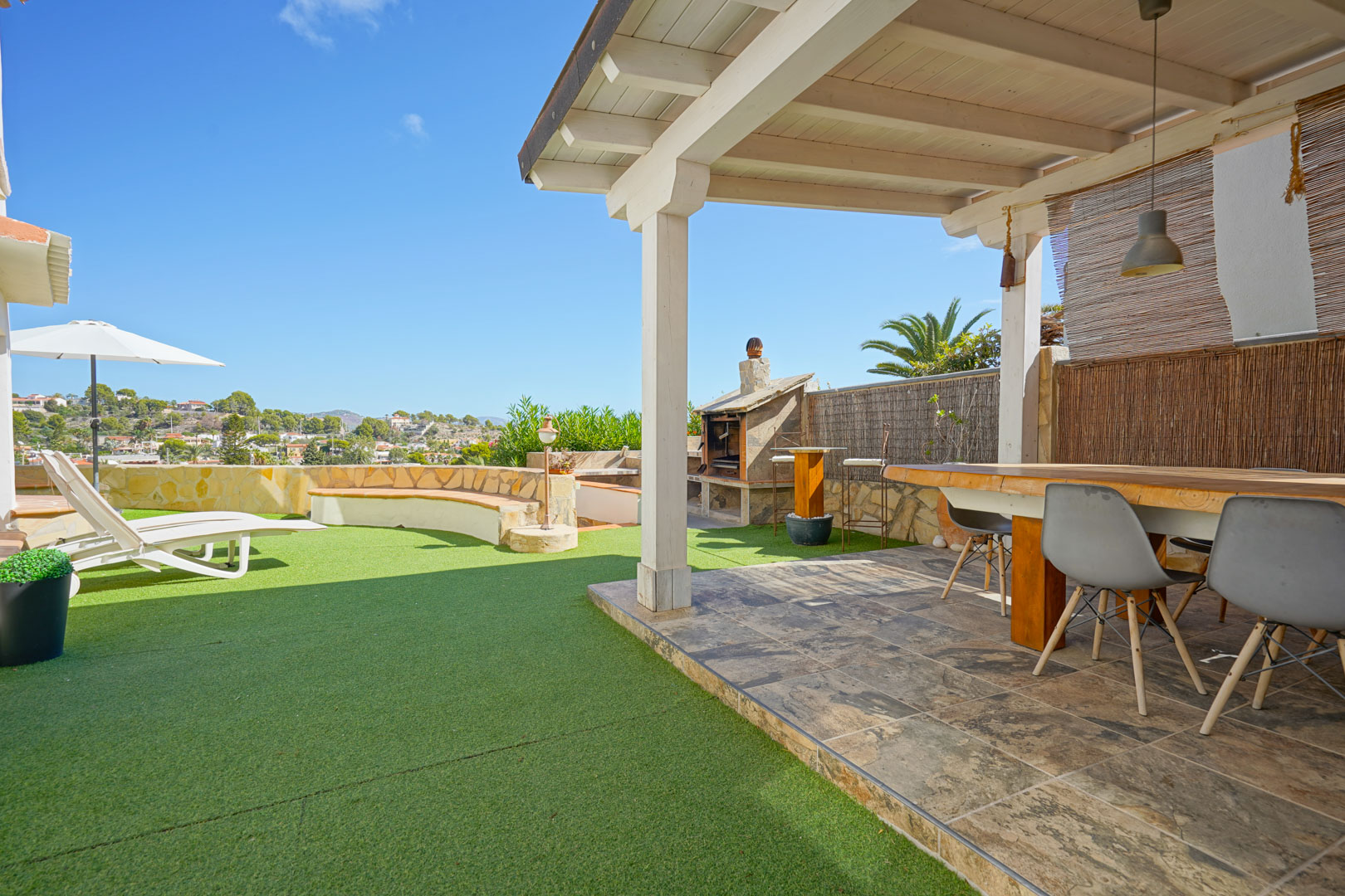 Terraza exterior con pérgola de madera blanca, zona de comedor, barbacoa de piedra y área de césped artificial con tumbonas y vistas despejadas.