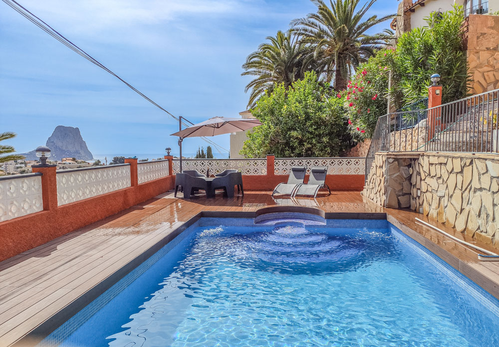 Terraza exterior con piscina de mosaico azul, suelo de tarima y vistas despejadas al Peñón de Ifach. Dispone de zona de comedor bajo sombrilla y tumbonas.