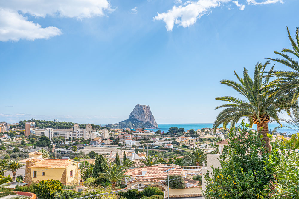 Vista panorámica desde la villa hacia el Peñón de Ifach y el mar Mediterráneo, enmarcada por palmeras y el paisaje urbano de Calpe bajo un cielo despejado.
