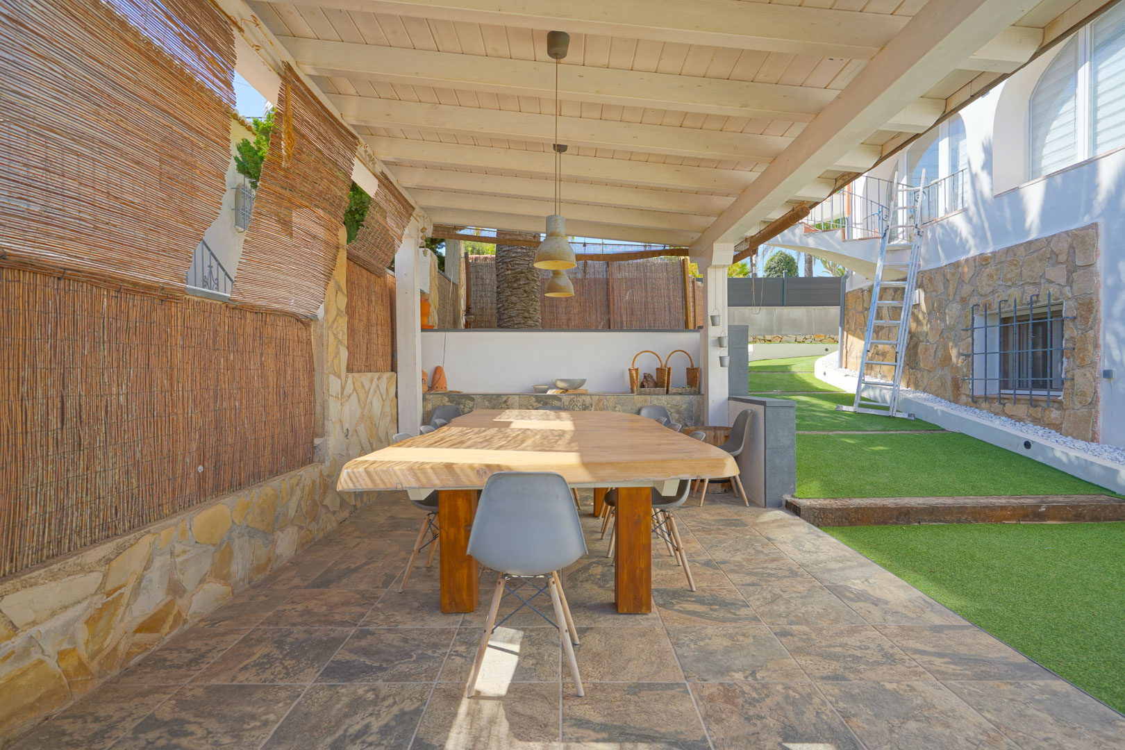Zona de comedor exterior bajo pérgola de madera blanca, con mesa de madera maciza y suelos cerámicos, rodeada de muros de piedra y césped artificial.