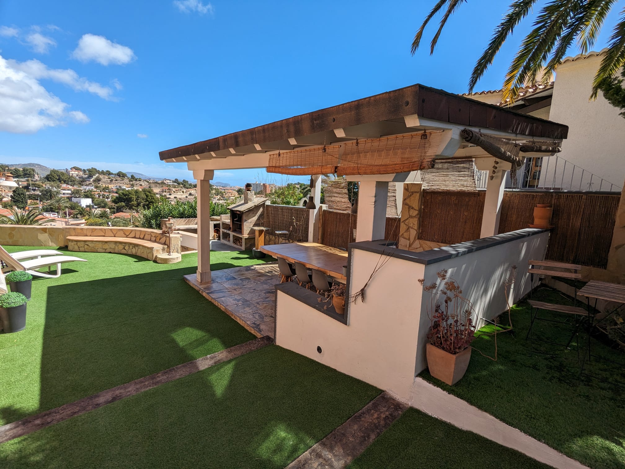 Zona de comedor exterior bajo pérgola de madera con mesa robusta, suelo cerámico y barbacoa de obra integrada, rodeada de césped artificial y vistas despejadas.