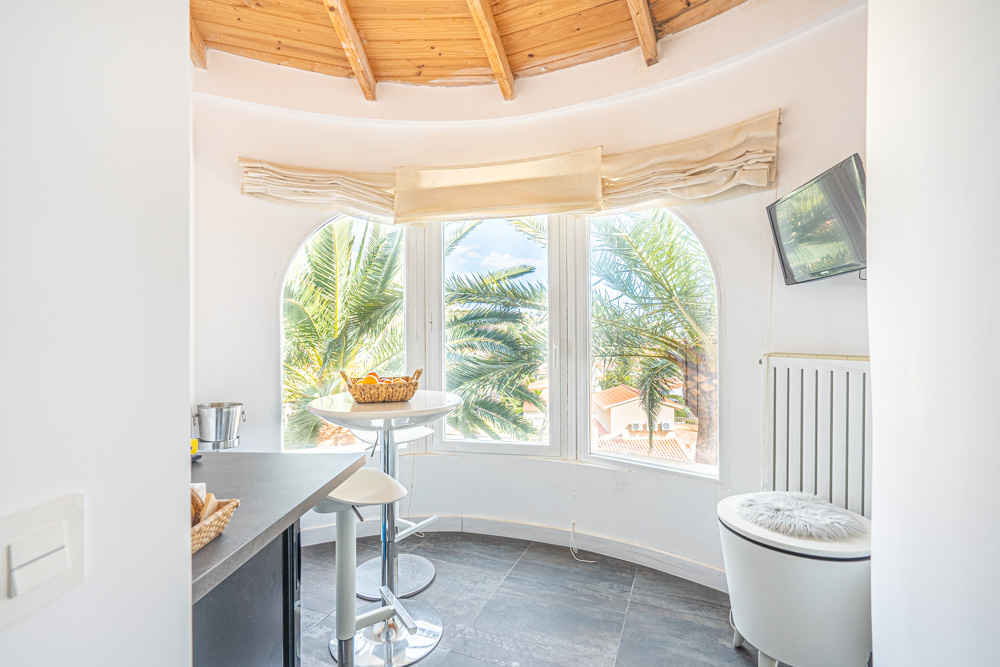 Zona de desayuno circular con techo de vigas de madera, ventanales panorámicos hacia las palmeras y mobiliario de bar integrado en un espacio luminoso.