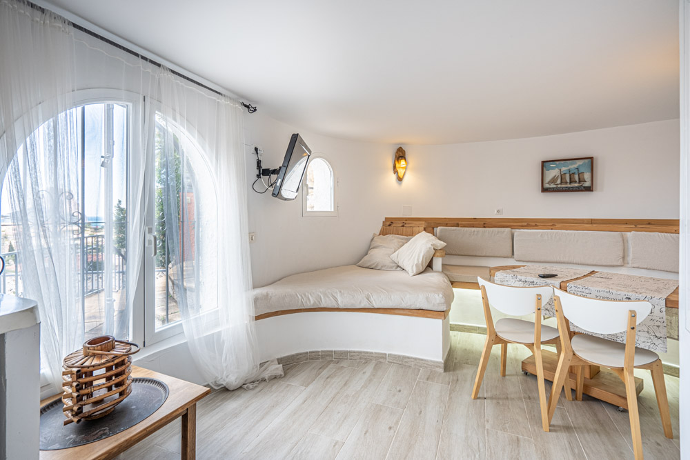 Zona de estar con banco de madera integrado, mesa de comedor y sillas blancas. Destaca el ventanal de arco con cortinas ligeras y suelos de gres efecto madera.