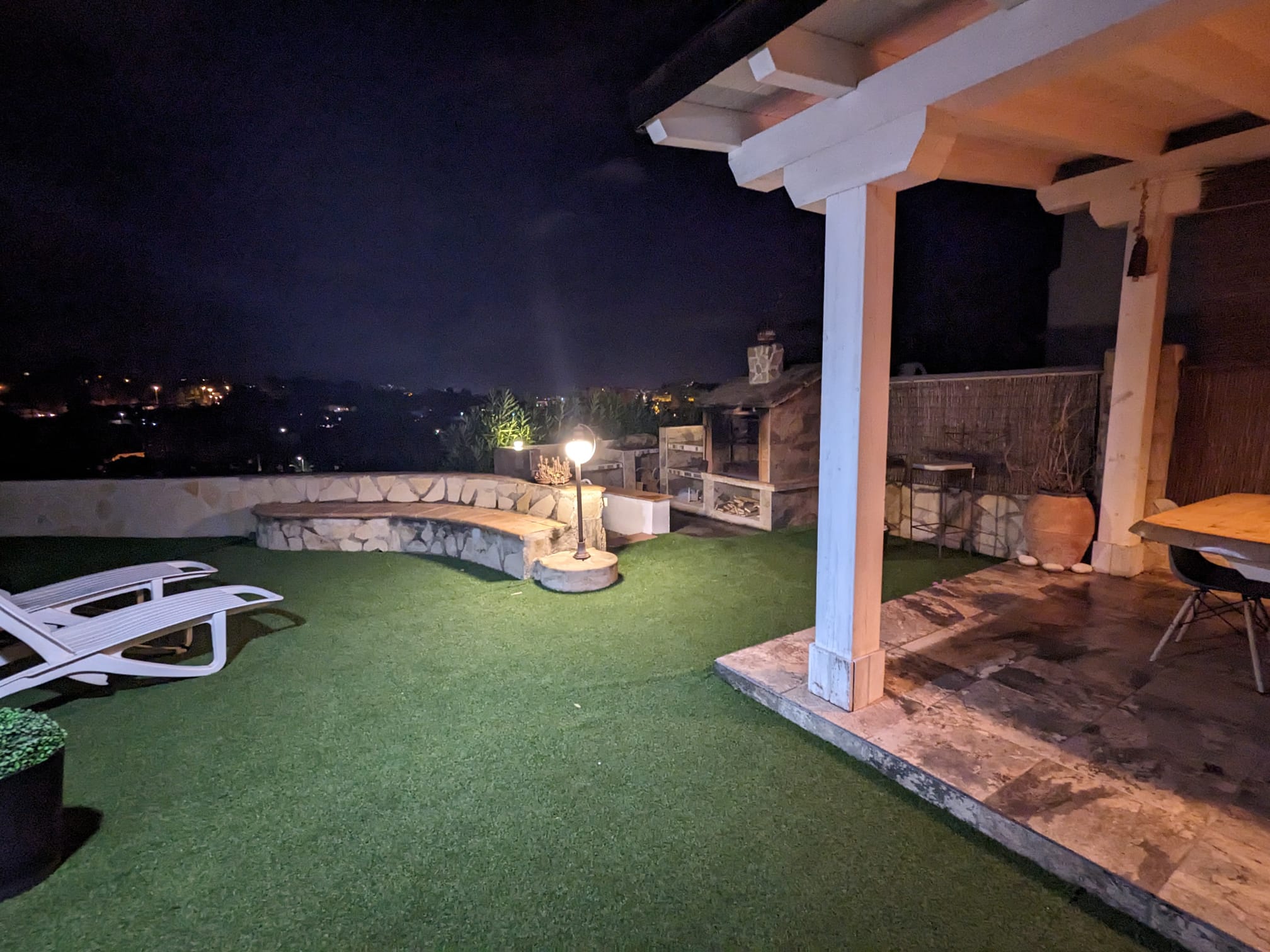 Zona exterior nocturna con barbacoa de piedra, banco curvo de mampostería y pérgola de madera iluminada junto a un área de césped artificial con tumbonas.