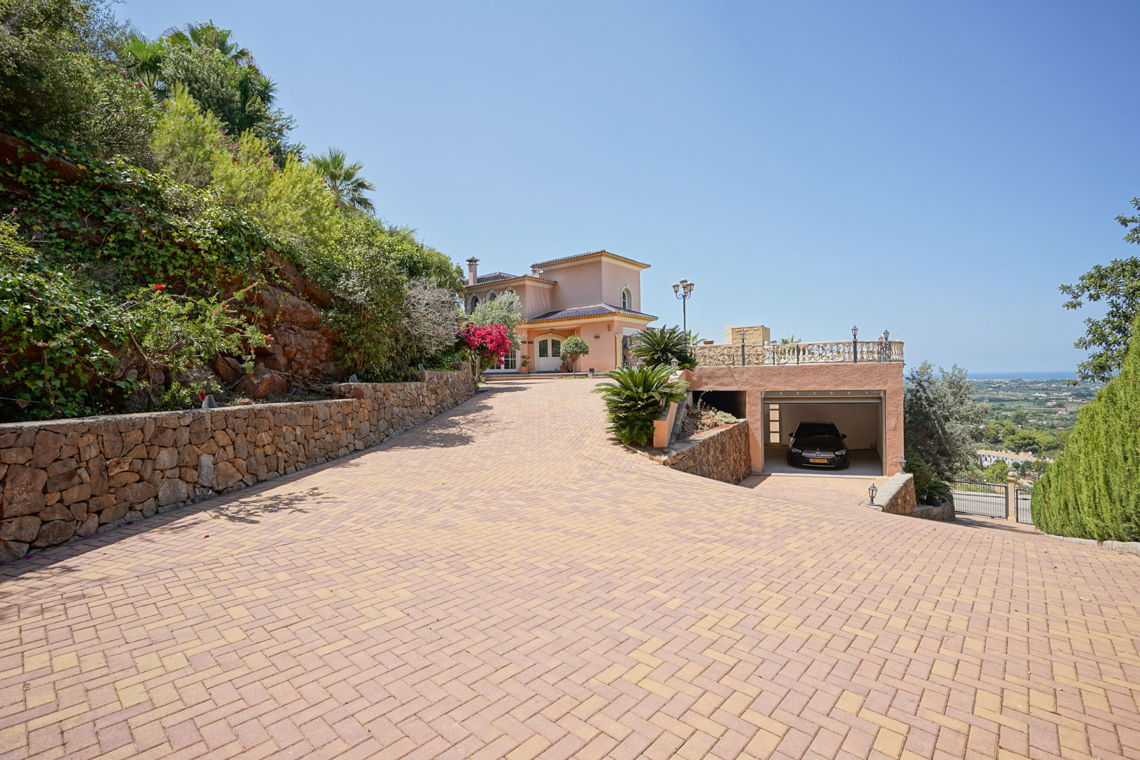 Amplio acceso pavimentado que conduce a la villa y al garaje integrado, flanqueado por muros de piedra natural, jardines mediterráneos y vistas panorámicas al mar.