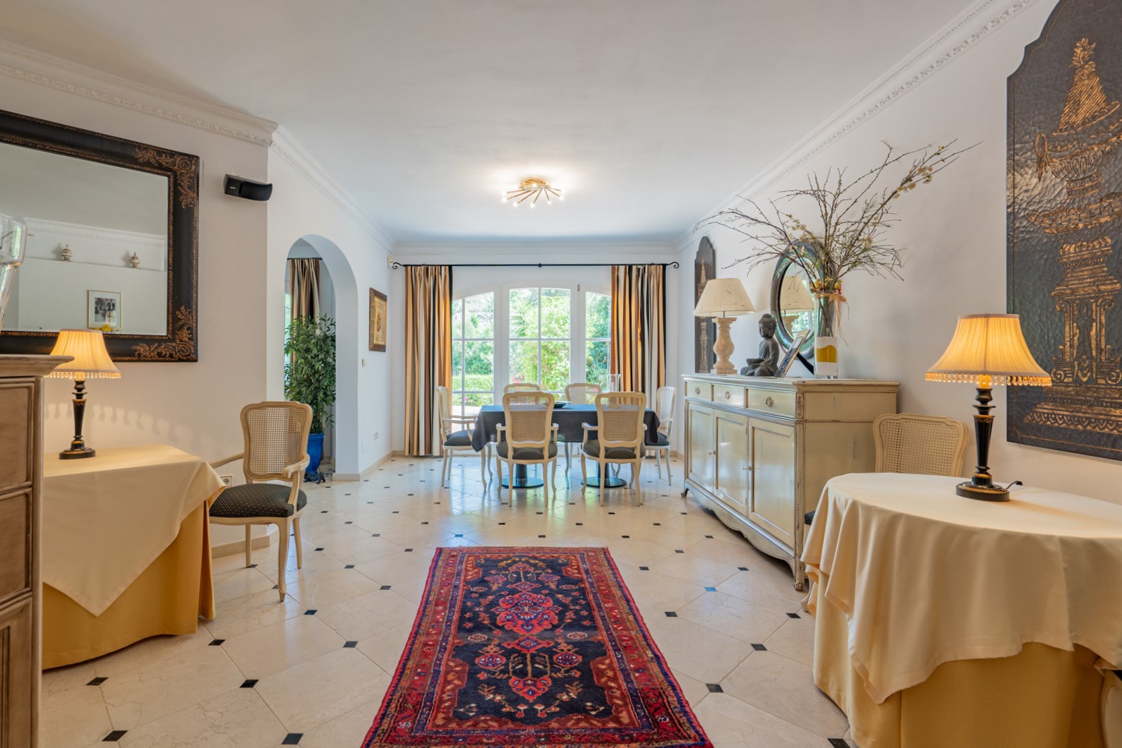 Amplio comedor con suelos de mármol y detalles en negro, mobiliario de estilo clásico y grandes ventanales de arco que conectan con el jardín exterior.