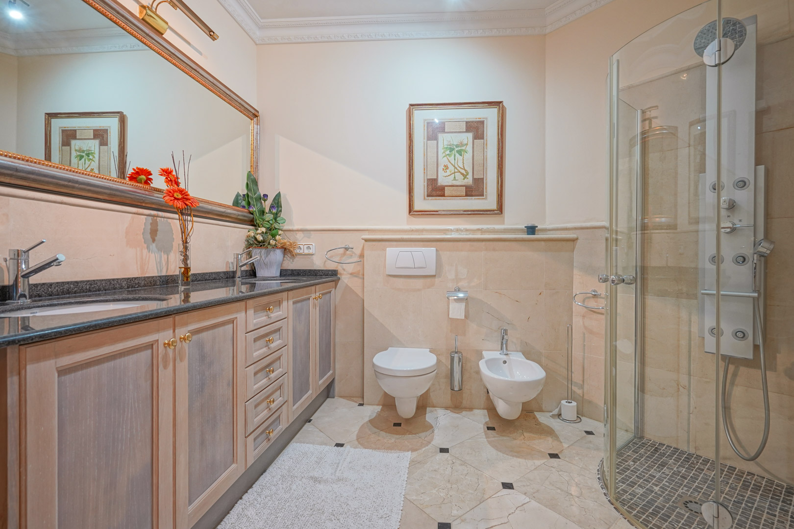 Baño con encimera de granito oscuro, lavabo doble, inodoro suspendido y una ducha con columna de hidromasaje y mampara de cristal sobre suelos de mármol.