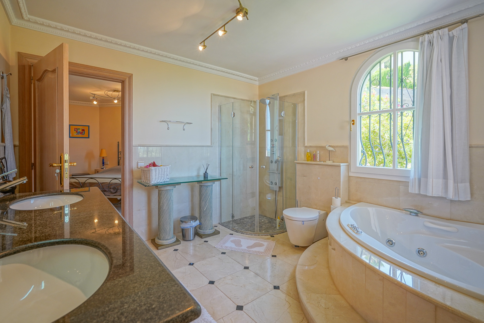 Baño principal con bañera de hidromasaje circular, ducha acristalada y tocador doble de granito. Cuenta con suelos de mármol y un ventanal de arco con vistas al jardín exterior.