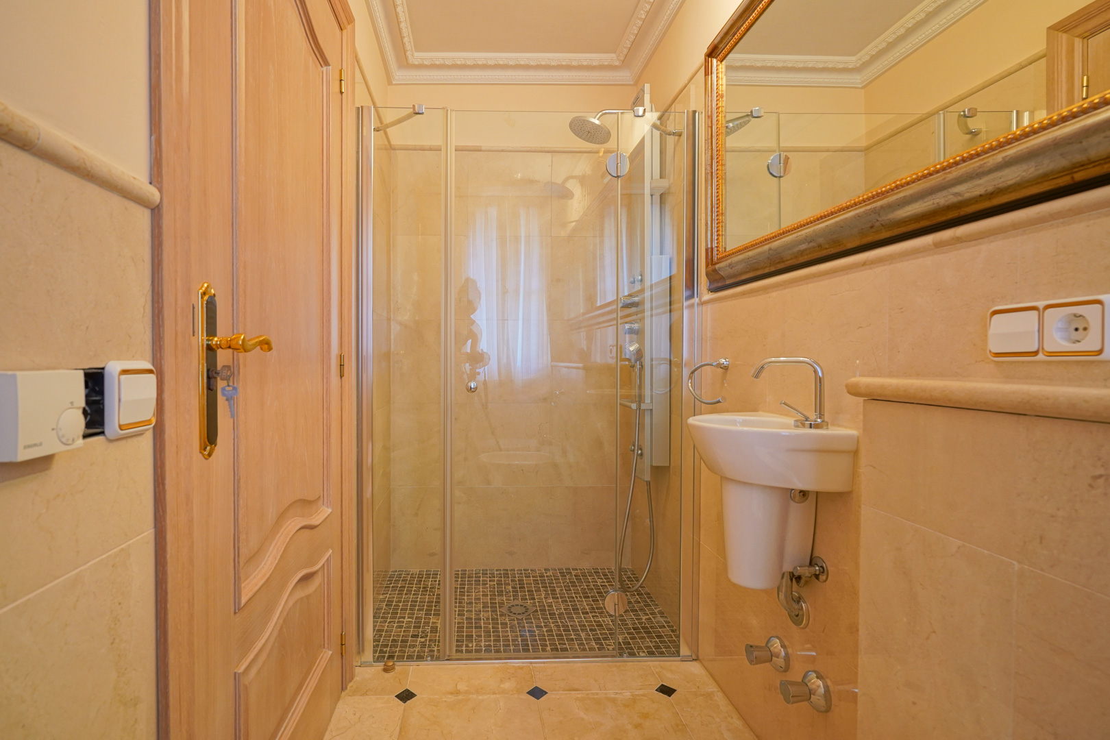 Baño revestido en mármol con ducha de obra, suelo de mosaico y mampara de cristal. Incluye un lavabo suspendido y espejo con marco dorado.
