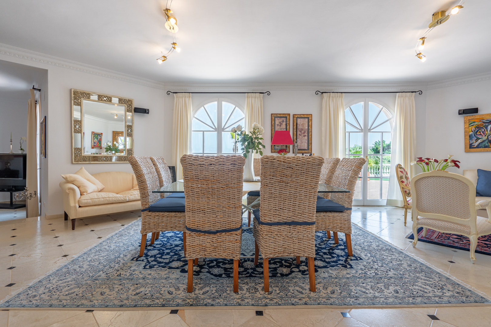 Comedor elegante con mesa de cristal y sillas de mimbre sobre alfombra clásica. Grandes ventanales de arco aportan luz natural y acceso directo a la terraza.