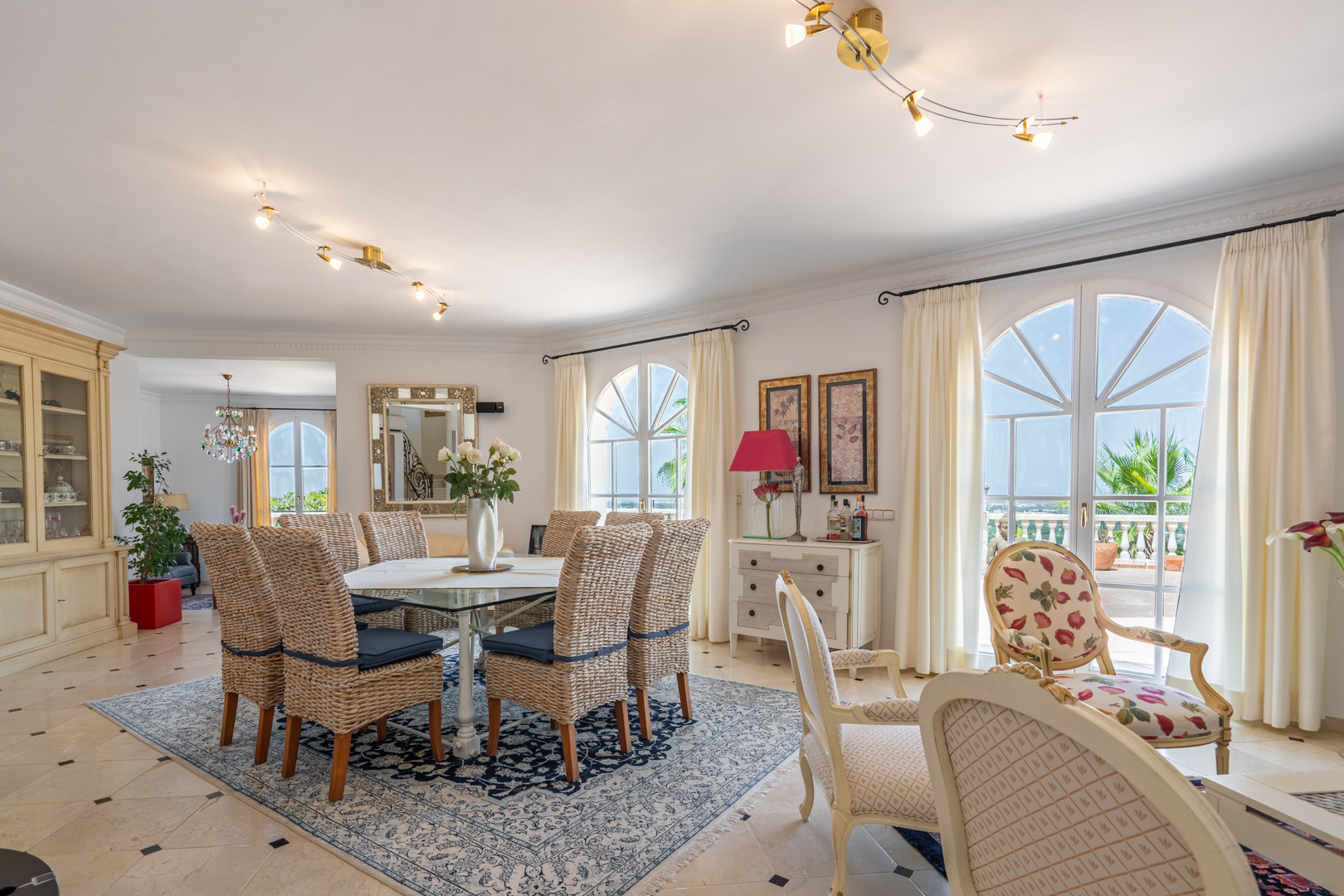 Comedor luminoso con sillas de mimbre y mesa de cristal sobre alfombra clásica, con acceso directo a la terraza a través de ventanales de medio punto.