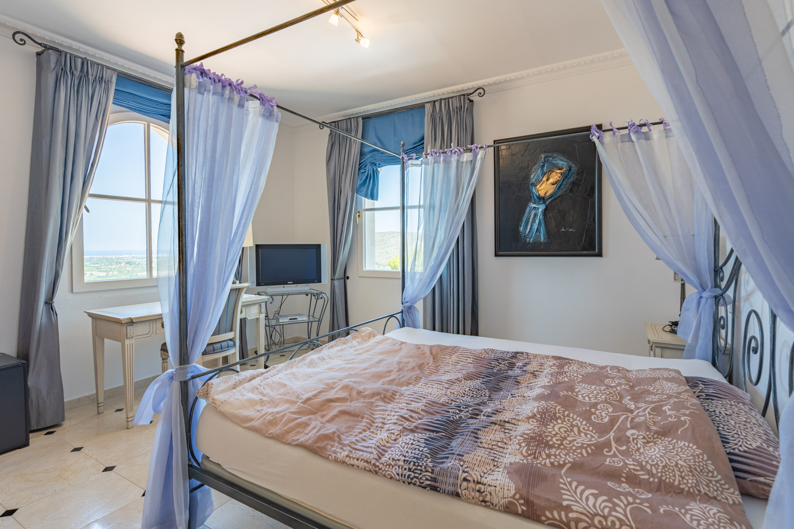 Dormitorio con cama de dosel metálica y cortinas ligeras, suelos de mármol con detalles en negro y escritorio junto a la ventana con vistas al valle.
