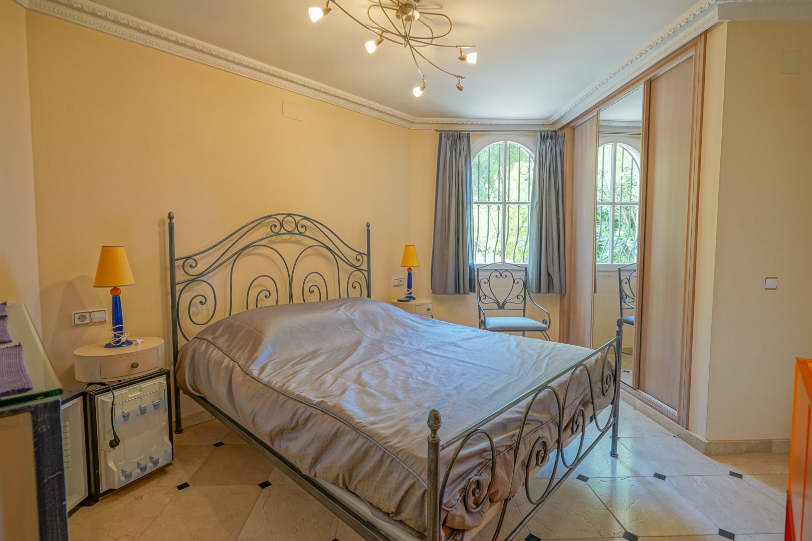 Dormitorio con cama de hierro forjado, suelos de mármol con detalles en negro, armarios empotrados y ventana de arco con vistas al exterior.