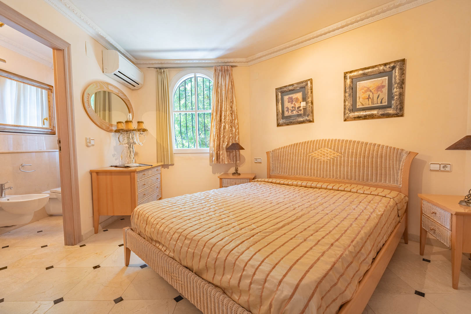 Dormitorio con cama de mimbre, suelos de mármol con detalles oscuros y baño en suite. Incluye aire acondicionado, molduras decorativas y ventana de arco con luz natural.