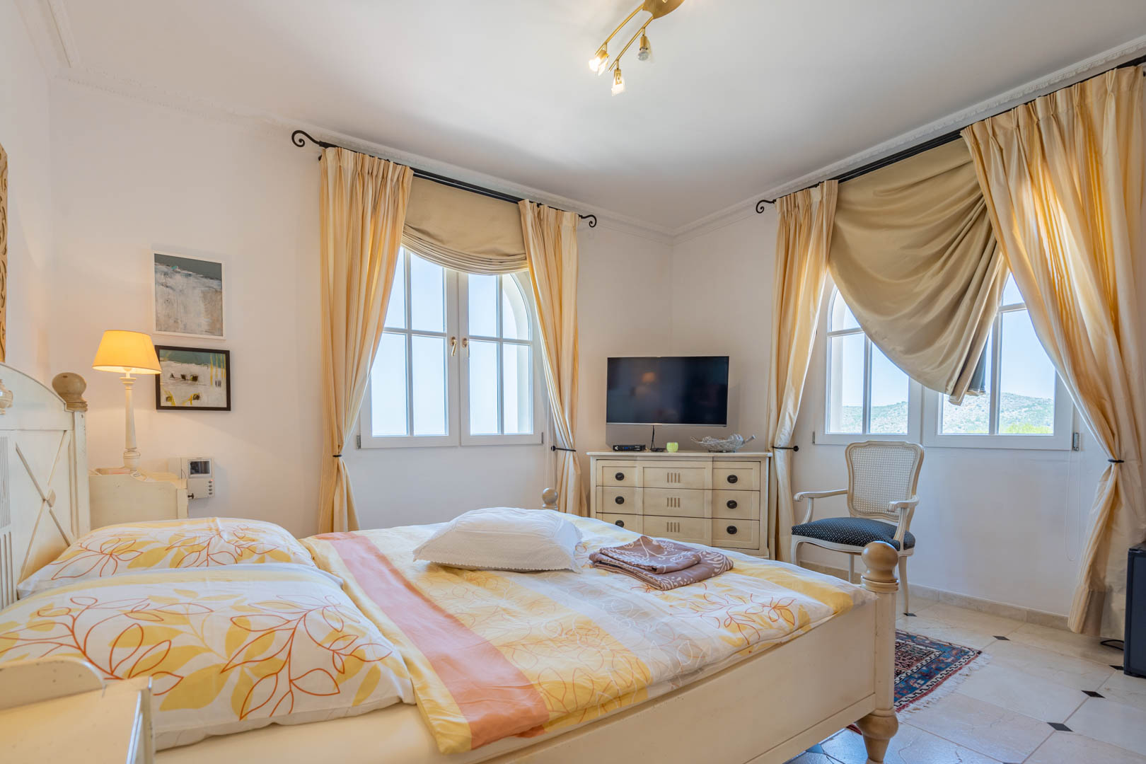 Dormitorio luminoso con mobiliario de madera blanca, ventanas de arco con cortinas crema y suelos cerámicos con detalles en contraste. Incluye zona de televisión y rincón de lectura.
