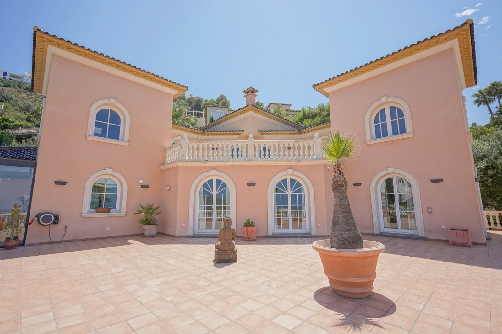 Fachada simétrica de tonos salmón con ventanales de arco, terraza superior con balaustrada blanca y un amplio patio de baldosas de barro con elementos decorativos.