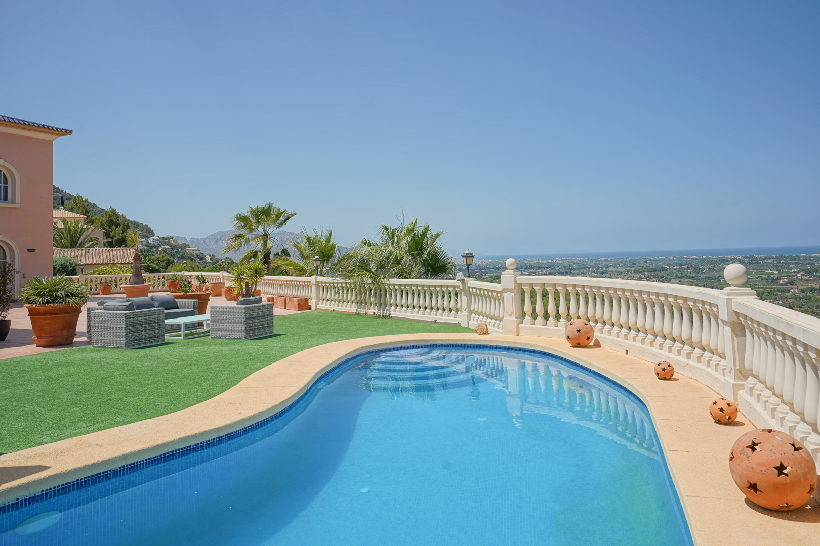 Piscina de forma irregular con vistas panorámicas al mar y al valle, rodeada de una balaustrada clásica, zona de césped artificial y mobiliario de exterior.