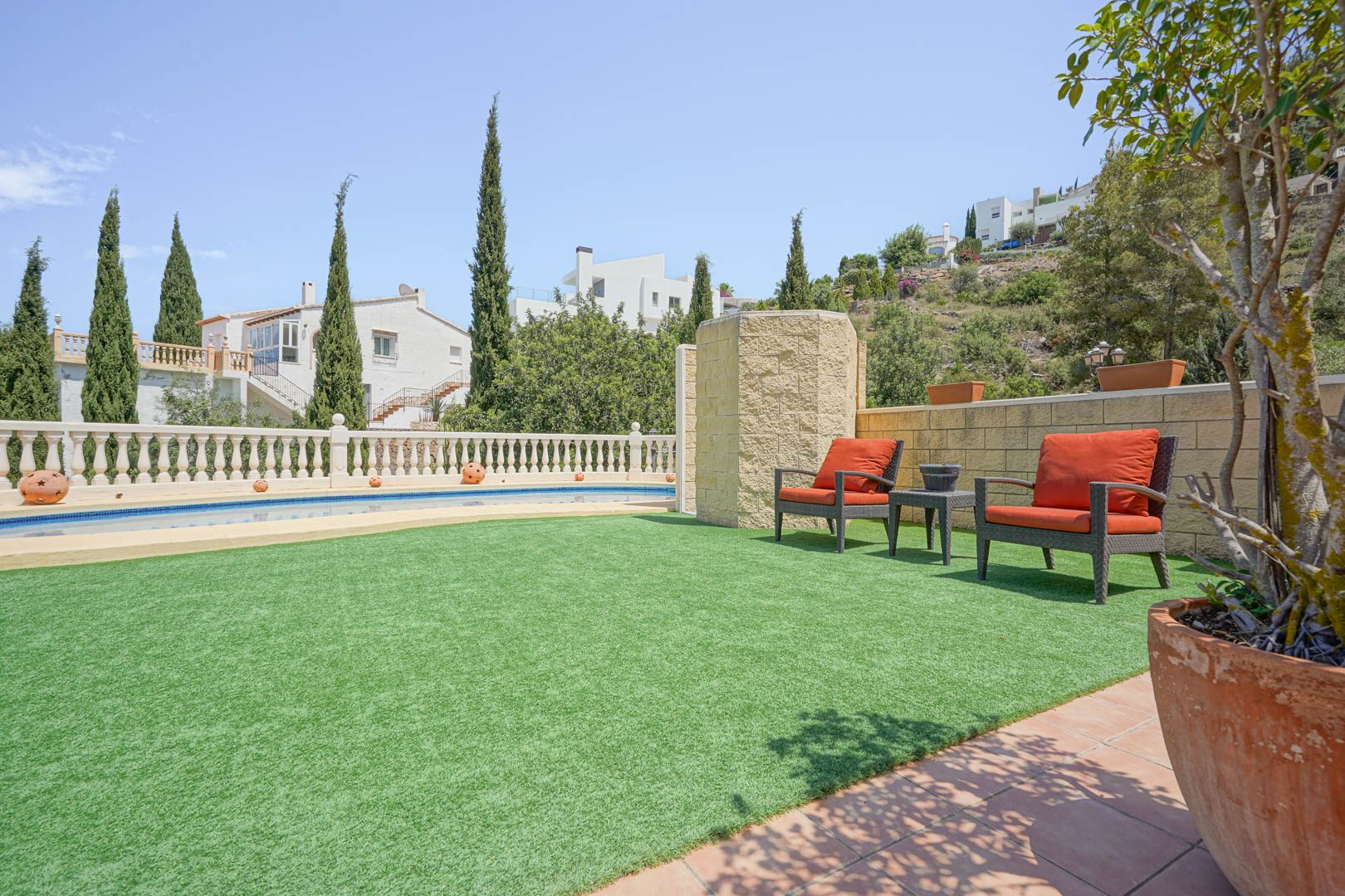Terraza exterior con césped artificial, zona de descanso con sillones naranjas y piscina privada delimitada por una balaustrada clásica y cipreses.