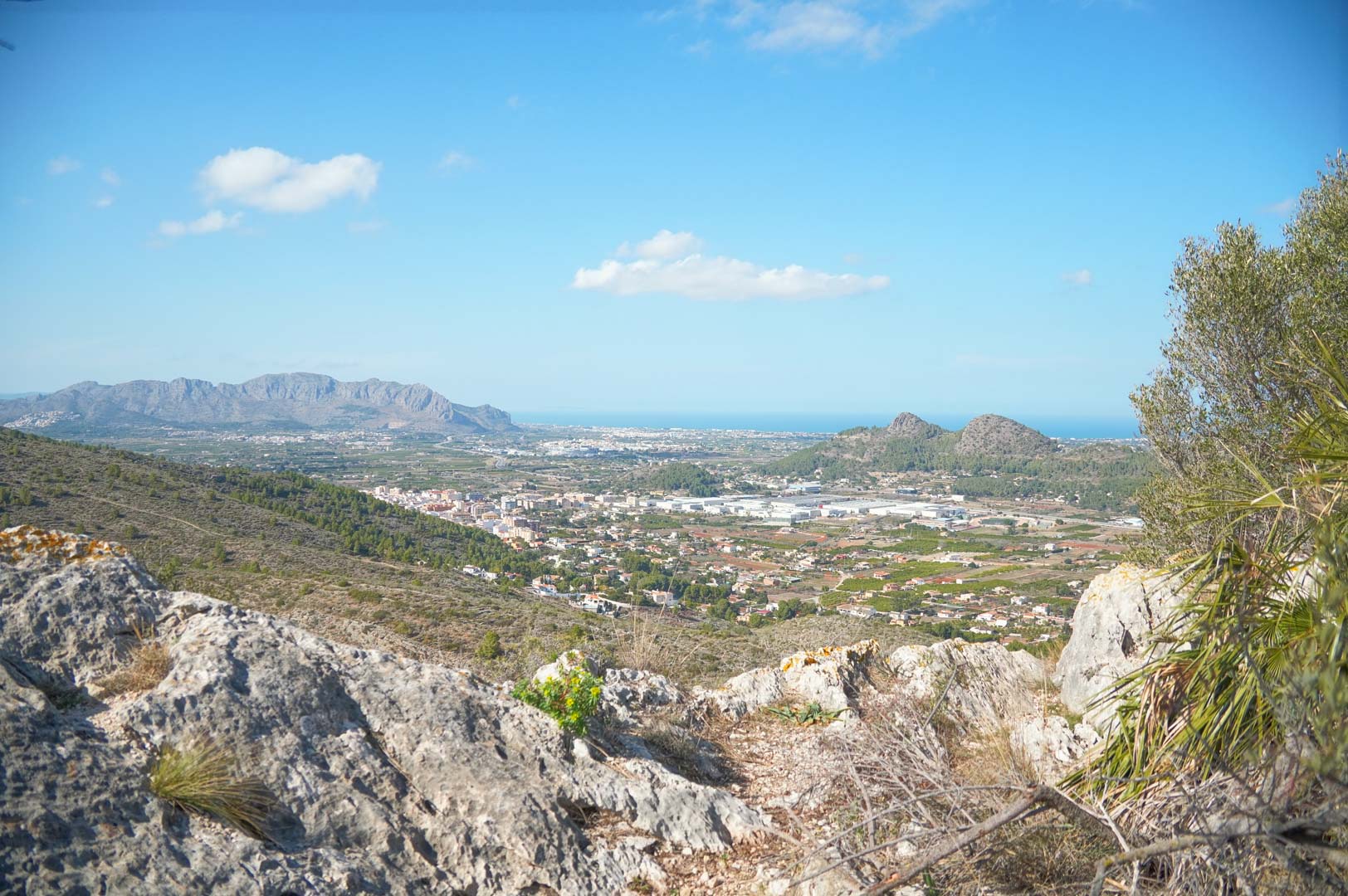 Vistas panorámicas desde la posición elevada de La Sella, abarcando el valle, las montañas circundantes y el mar Mediterráneo bajo un cielo despejado.