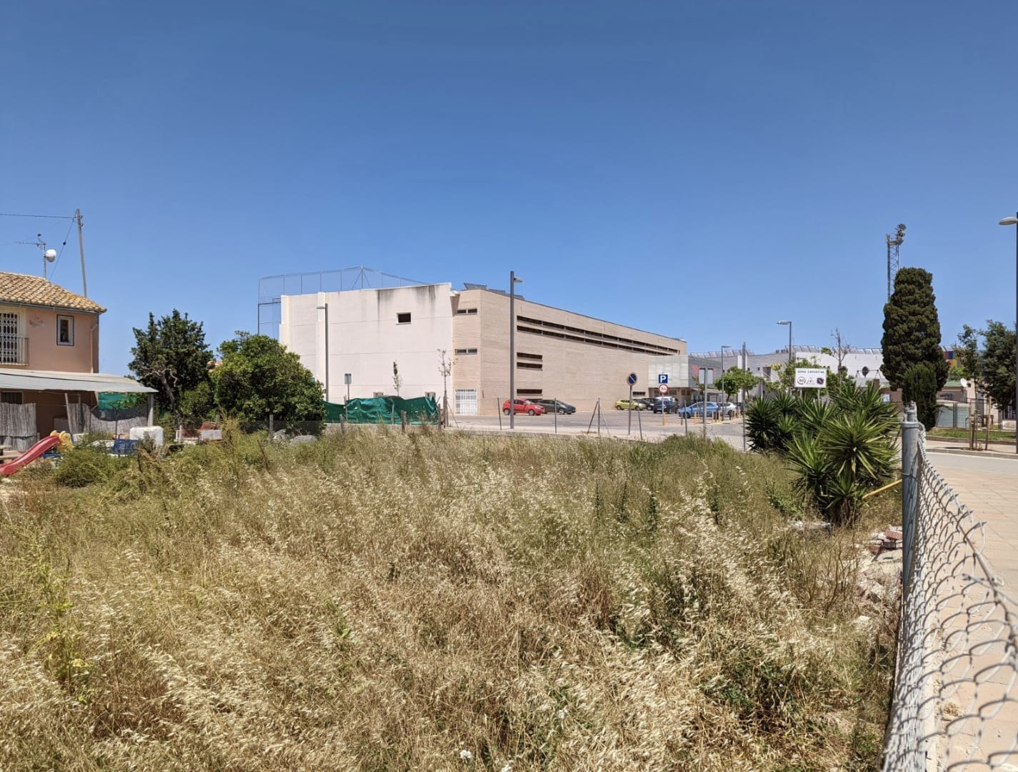 Parcela urbana llana con vegetación natural, situada frente a un centro deportivo moderno y zona de aparcamiento bajo un cielo despejado.