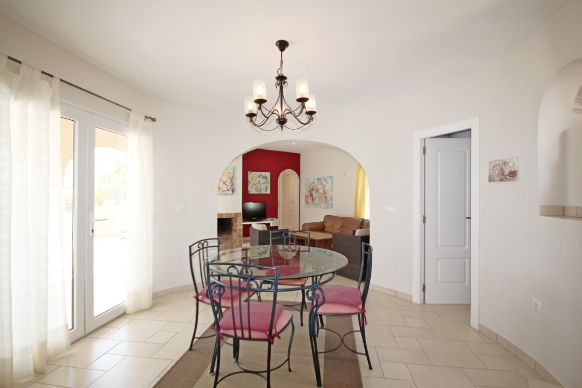 Comedor con mesa circular de cristal y sillas de forja, iluminado por una lámpara clásica y conectado al salón con chimenea a través de un arco arquitectónico.