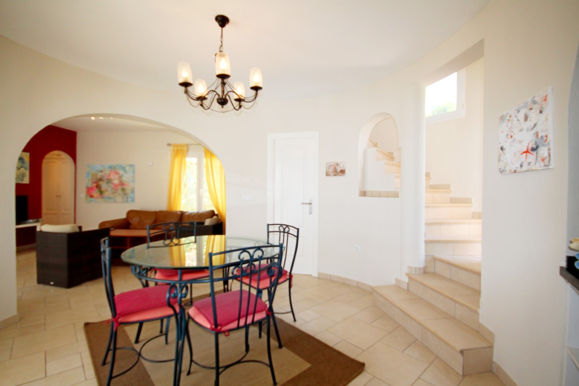Comedor con mesa de cristal y sillas de forja, conectado al salón mediante un arco y situado junto a la escalera de baldosas claras que sube a la planta superior.
