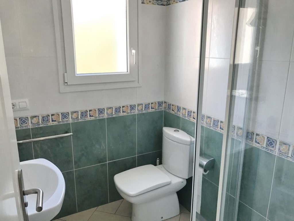 Cuarto de baño con azulejos verdes y blancos, inodoro de porcelana, lavabo y ducha con mampara de cristal. Una ventana proporciona luz natural al espacio.