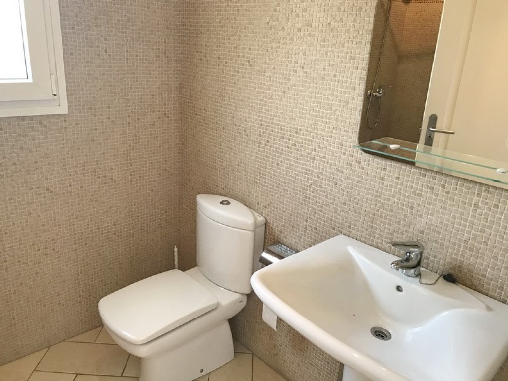 Cuarto de baño con paredes de mosaico beige, lavabo de cerámica blanca y espejo con estante de cristal. Suelos de gres en tonos neutros y ventana lateral para luz natural.