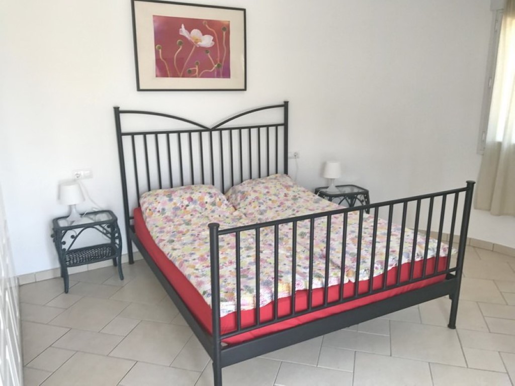 Dormitorio doble con estructura de cama de hierro forjado negro, mesitas de noche de mimbre a juego y suelos de baldosas cerámicas claras.