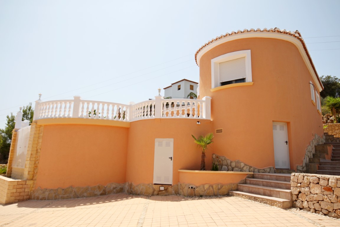 Fachada exterior de estilo mediterráneo con torre circular en tono terracota, balustrada blanca y patio pavimentado con escaleras de piedra.