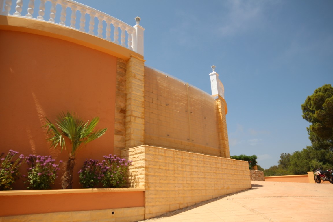 Muro exterior de piedra tosca y pintura terracota con paneles de cristal protectores, jardineras con palmeras pequeñas y balaustrada blanca superior.