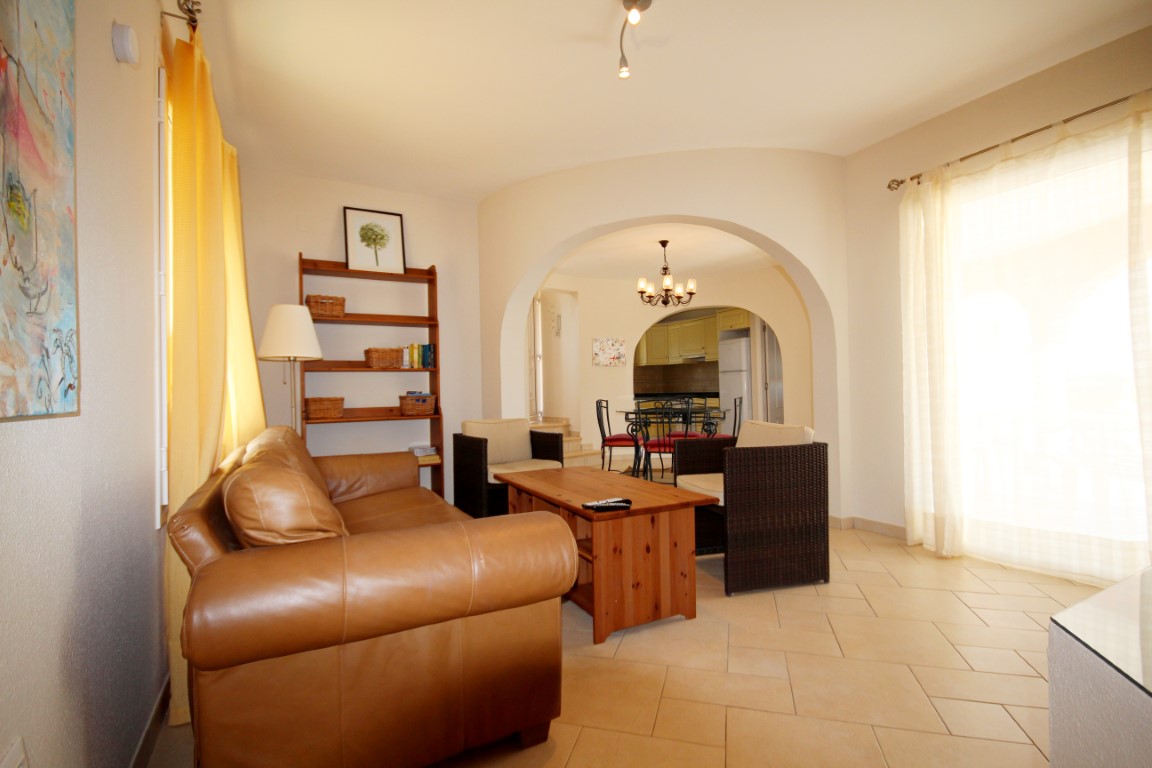 Salón con sofá de cuero, mesa de centro de madera y sillones de mimbre. Un arco arquitectónico comunica la estancia con el comedor y la cocina al fondo.