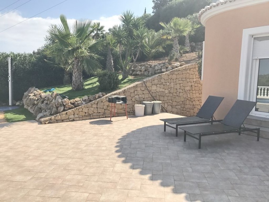 Terraza pavimentada con tumbonas y barbacoa portátil, delimitada por un muro de piedra natural y un jardín elevado con palmeras de estilo mediterráneo.