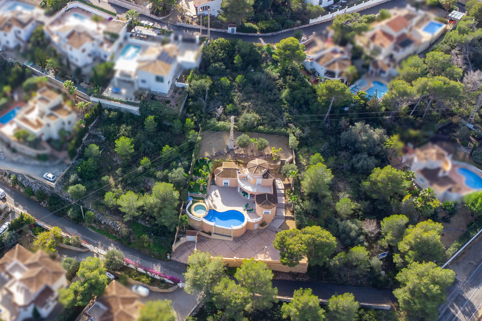 Vista aérea de la villa de estilo mediterráneo con piscina de forma orgánica, jacuzzi circular y amplias terrazas pavimentadas rodeadas de una densa zona boscosa.