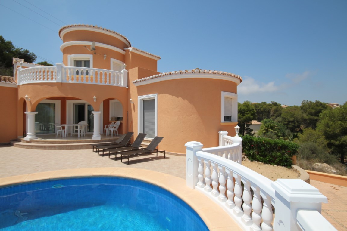 Vista exterior con fachada curva en tono terracota, balaustradas blancas y piscina privada. La terraza pavimentada incluye un porche cubierto con columnas y tumbonas.