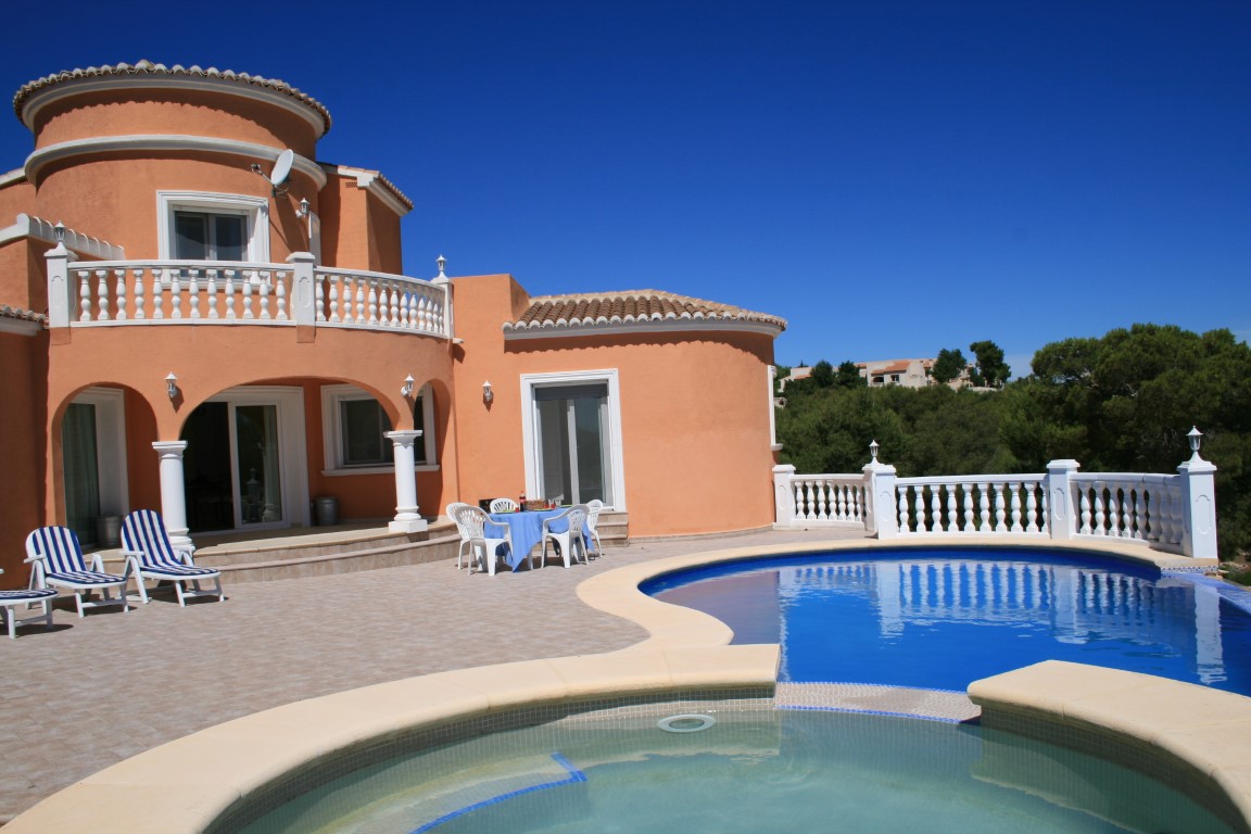 Vista exterior de una villa mediterránea con piscina de forma irregular, jacuzzi integrado y amplia terraza de baldosas con balaustradas blancas y arcos.